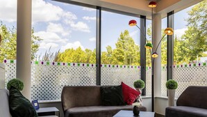 Un salon moderne comprenant un canapé, deux fauteuils, une table basse et un vase décoratif. De grandes fenêtres offrent une vue sur les arbres et une clôture.