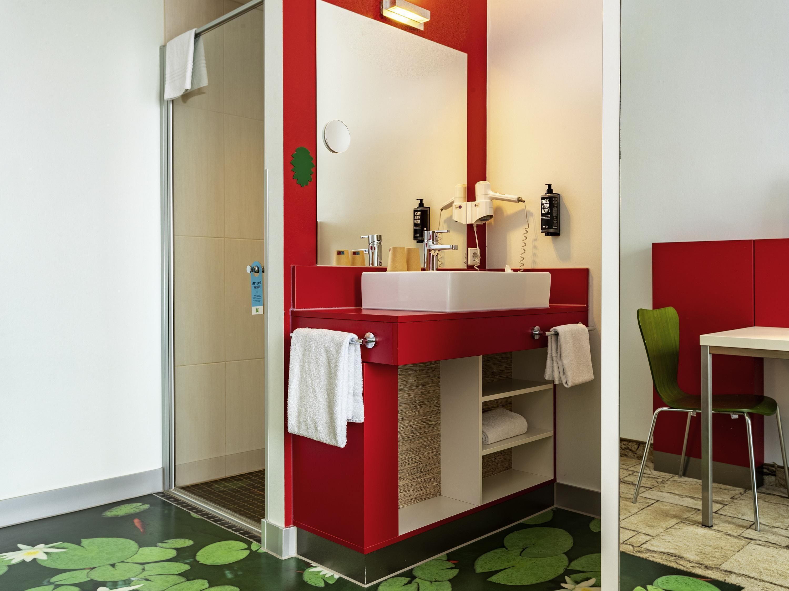 Un baño con tocador rojo, lavamanos blanco, espejo, toallero y zona de ducha.