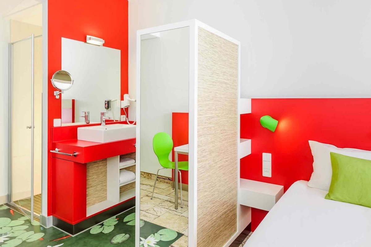 Une chambre d’hôtel aux couleurs rouge et blanc, avec une salle de bain séparée, une chaise verte et un lit agrémenté d’un oreiller vert.