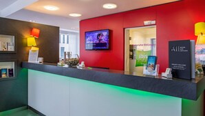 Un hall d’accueil d’hôtel avec un mur d’accent rouge, un téléviseur fixé au-dessus du comptoir et un panneau indiquant « Karlsruhe Ettlingen ».