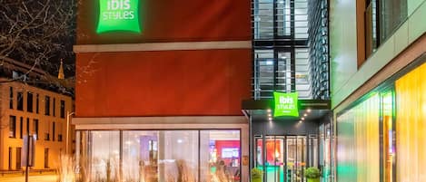 Un edificio moderno con un llamativo letrero de 'ibis Styles', grandes ventanales y una zona de entrada bien iluminada.
