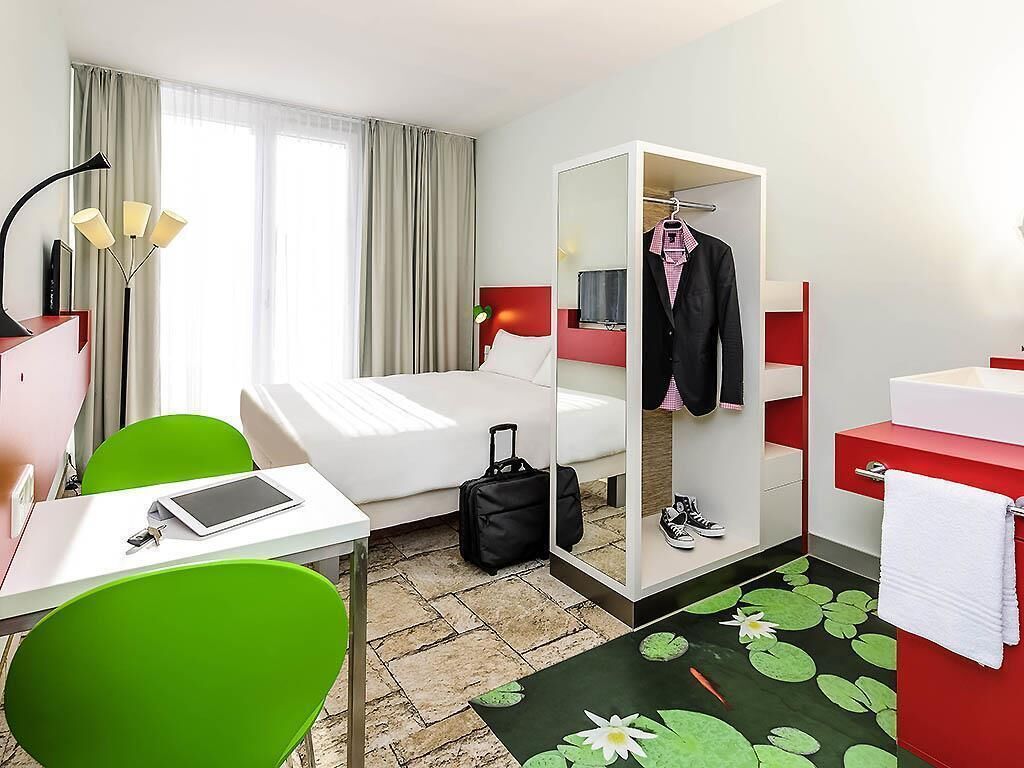 Une chambre d’hôtel avec un lit, un bureau doté d’une tablette, une chaise, une armoire, une valise, une salle de bain avec un lavabo rouge et une fenêtre avec des rideaux.