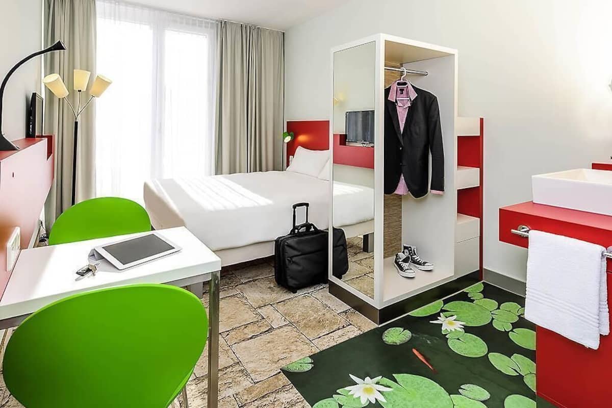 Une chambre d’hôtel avec un lit, un bureau doté d’une tablette, une chaise, une armoire, une valise, une salle de bain avec un lavabo rouge et une fenêtre avec des rideaux.