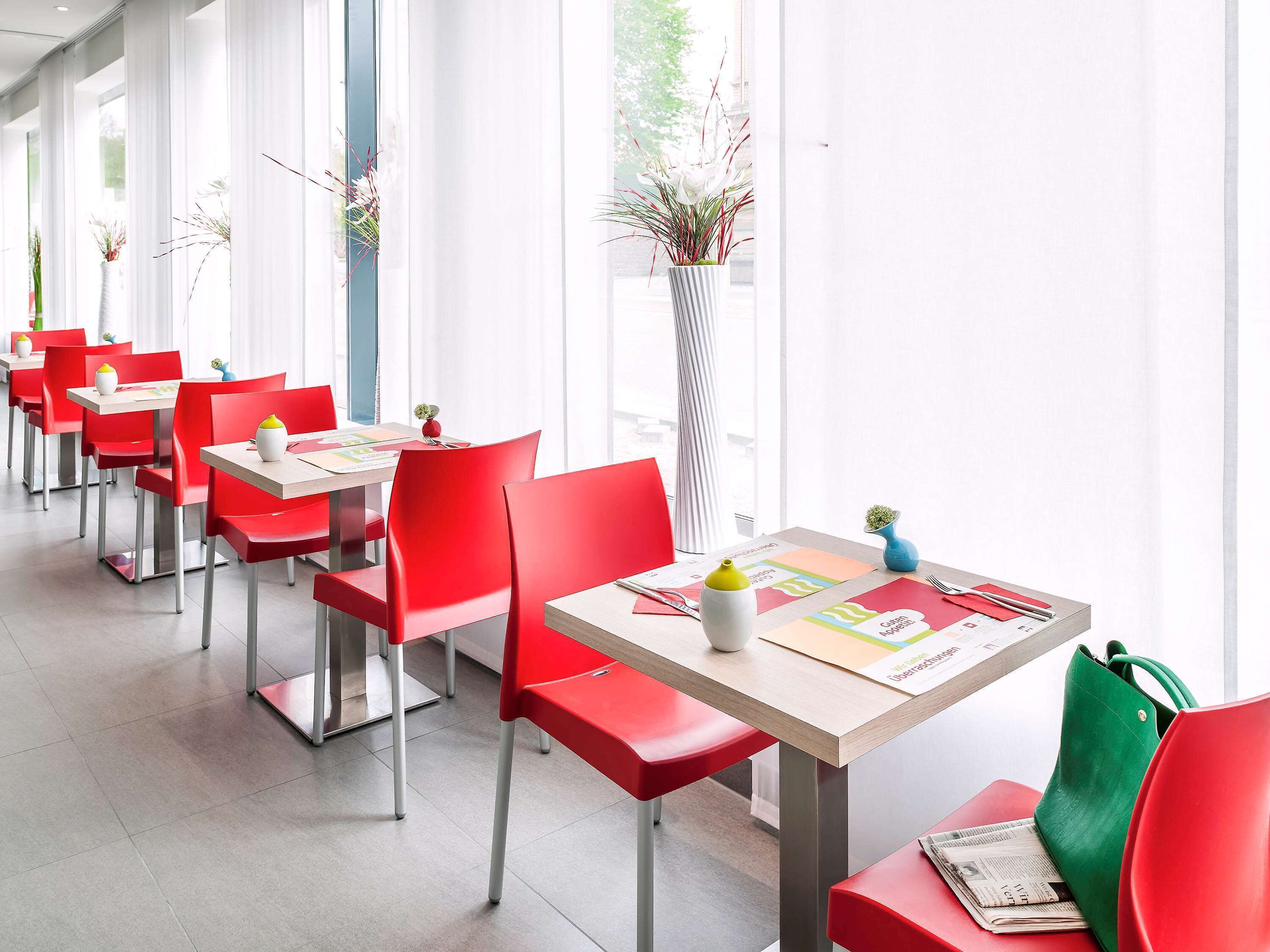 Un restaurante moderno con sillas rojas y mesas blancas, ventanas amplias y plantas de interior.