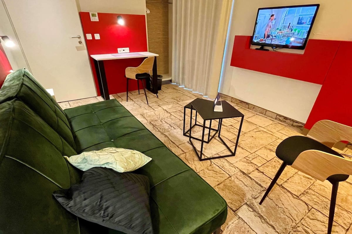 Een groene bank met een kussen, een klein zwart tafeltje, een stoel en een aan de muur bevestigde televisie.