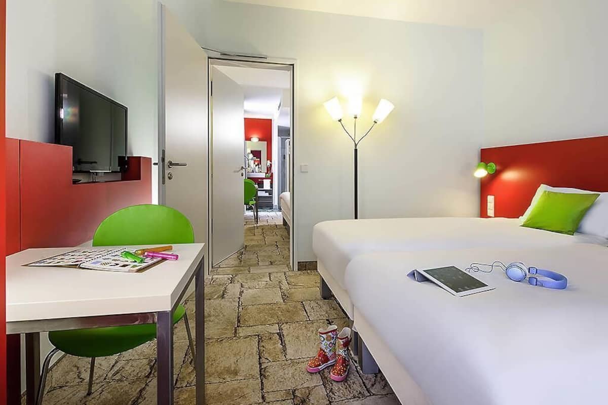 Een hotelkamer met een bed, een bureau met stoel, een televisie, een lamp en een deur die naar een andere kamer leidt.
