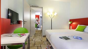 A hotel room with a bed, a desk with a chair, a TV, a lamp, and a door leading to another room.