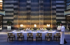 A dining area with a long table set for a meal, a waiter, and a decorative wall with a fire feature. - Bengaluru Marriott Hotel Whitefield (Bengaluru)
