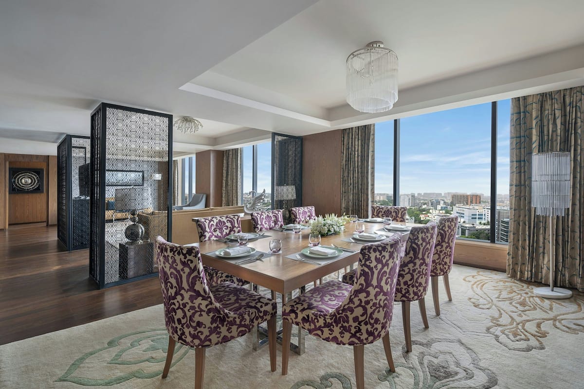A dining area with a large table set for six, surrounded by patterned chairs, a chandelier, and a view of the city.