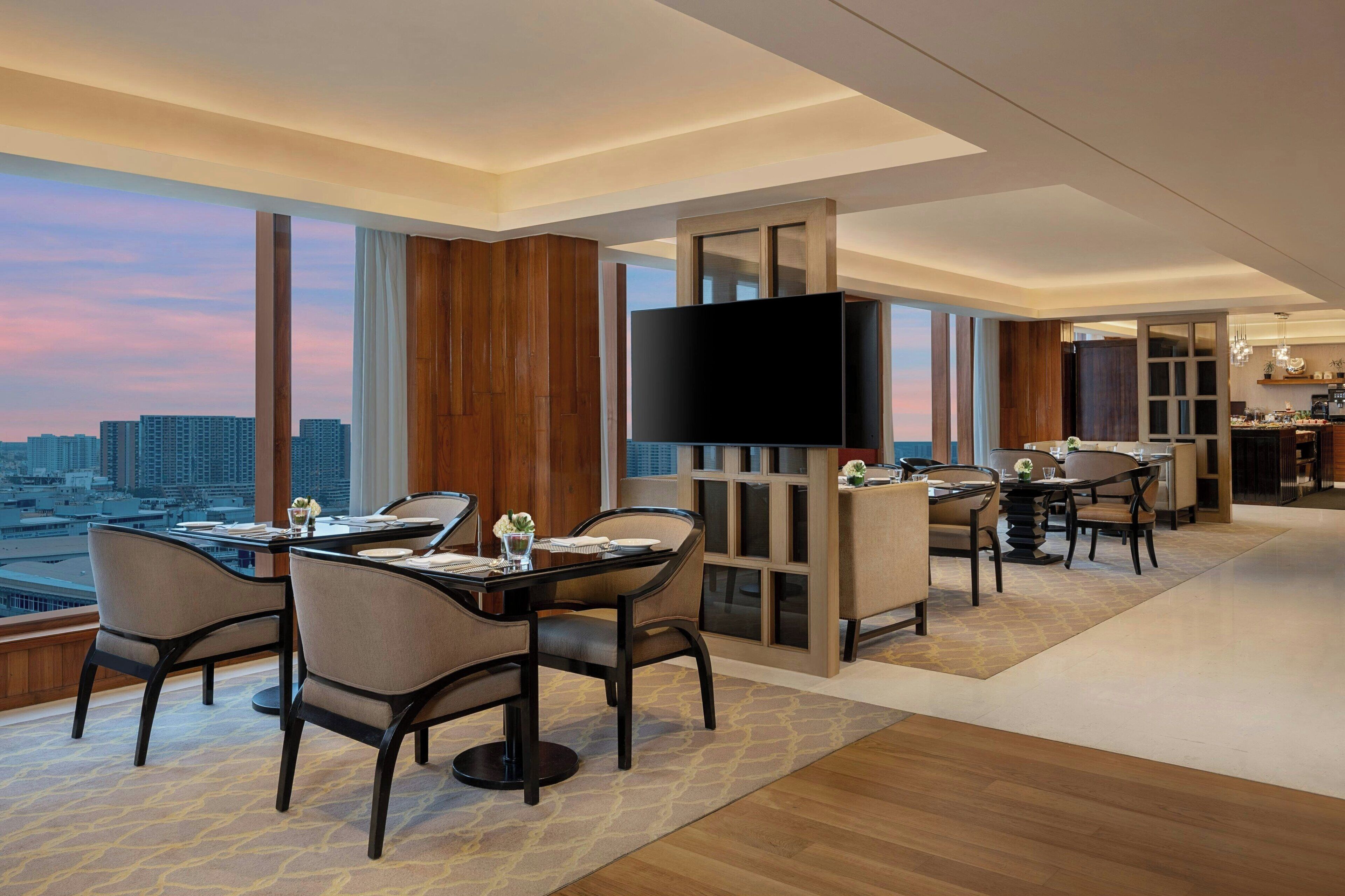 A modern hotel room with a dining area, a large TV, and a city view.