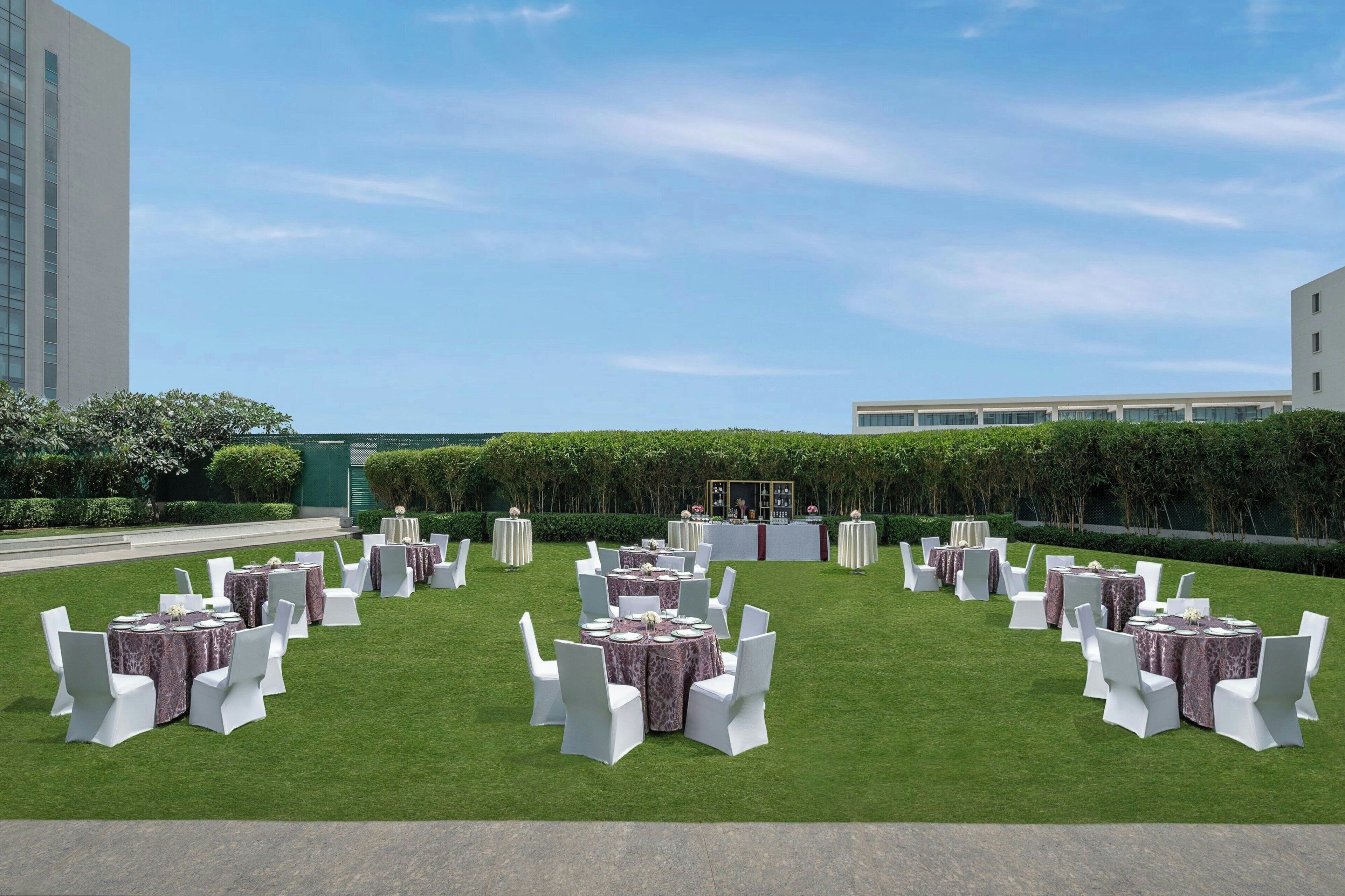 Un espacio para eventos al aire libre con mesas redondas y sillas blancas, rodeado de un césped cuidado y setos.