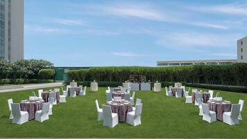 Un espacio para eventos al aire libre con mesas redondas y sillas blancas, rodeado de un césped cuidado y setos.
