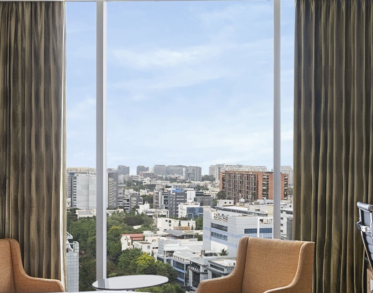 A hotel room with a large window, two chairs, a small table, and a view of a cityscape.