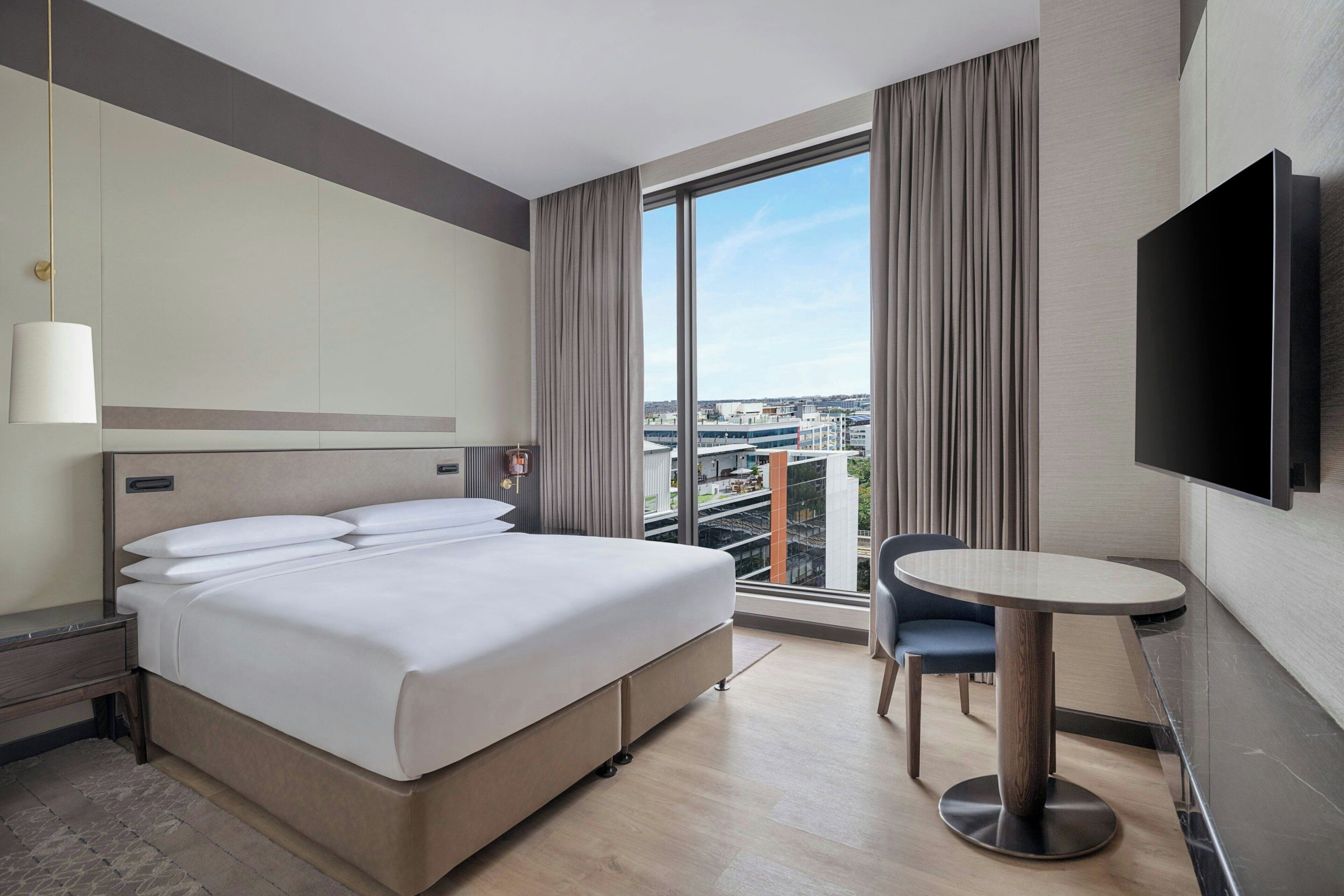 Een hotelkamer met een bed, een klein tafeltje, een stoel, een televisie en een groot raam met gordijnen.