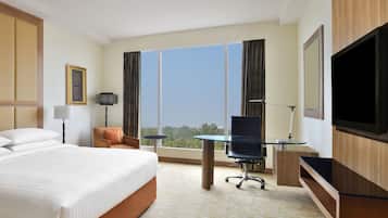 A hotel room with a large bed, a desk with a chair, a television, and a large window offering a city view.