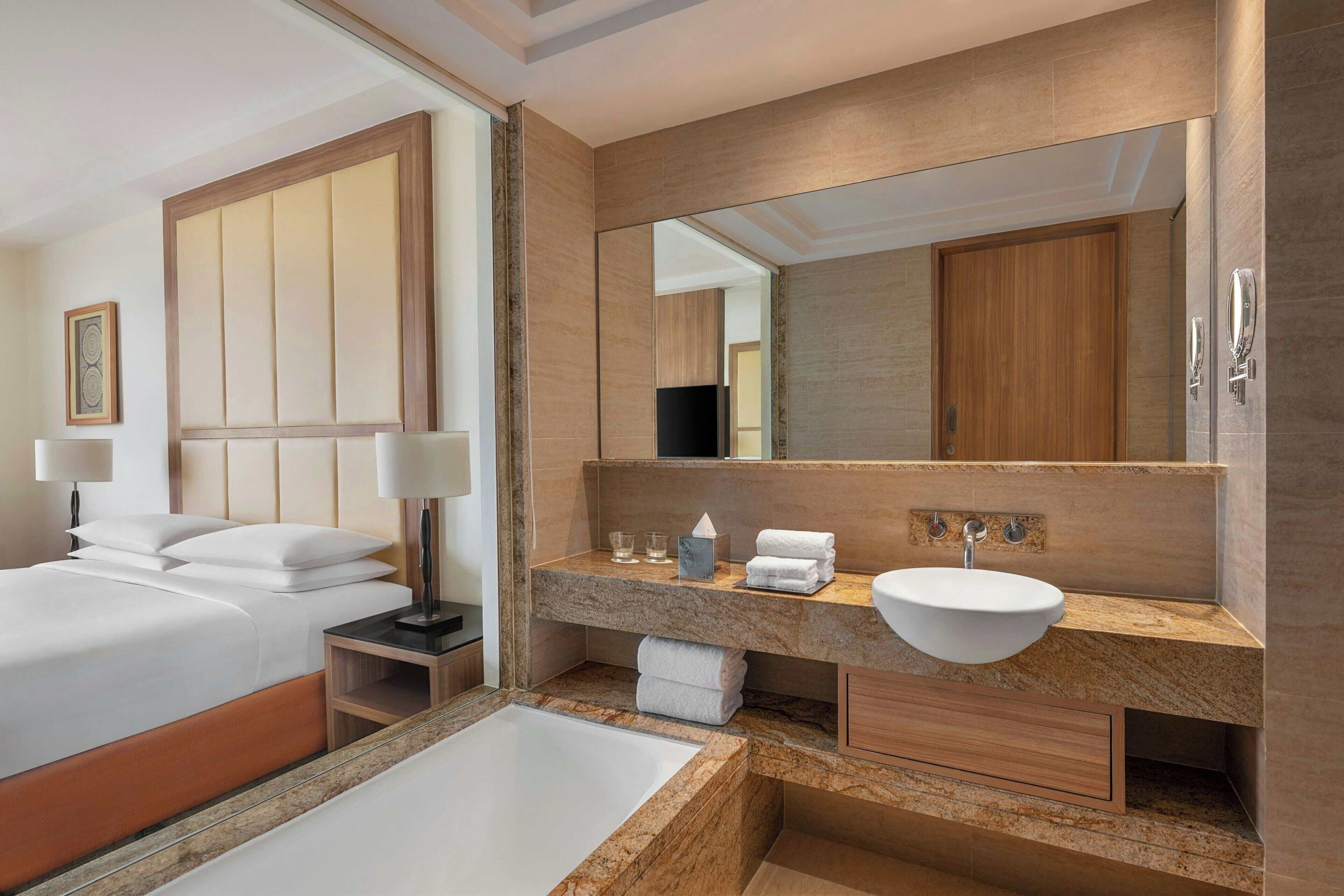 A modern hotel room with a large bed, a bathroom with a marble vanity, a large mirror, and a bathtub.