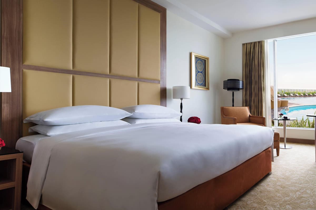 A hotel room with a large bed, a wooden headboard, a desk with a lamp, a chair, and a view of a pool.