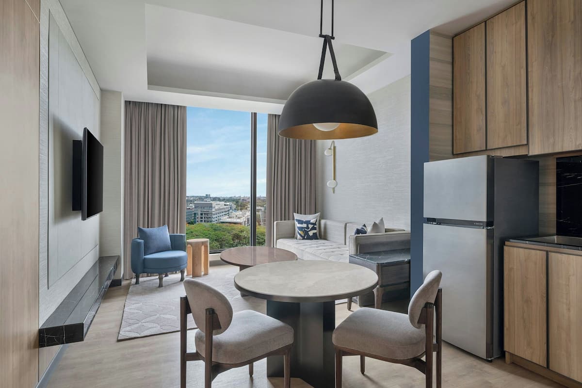 A modern kitchen with a dining area, a large window with a city view, and a flat-screen TV mounted on the wall.