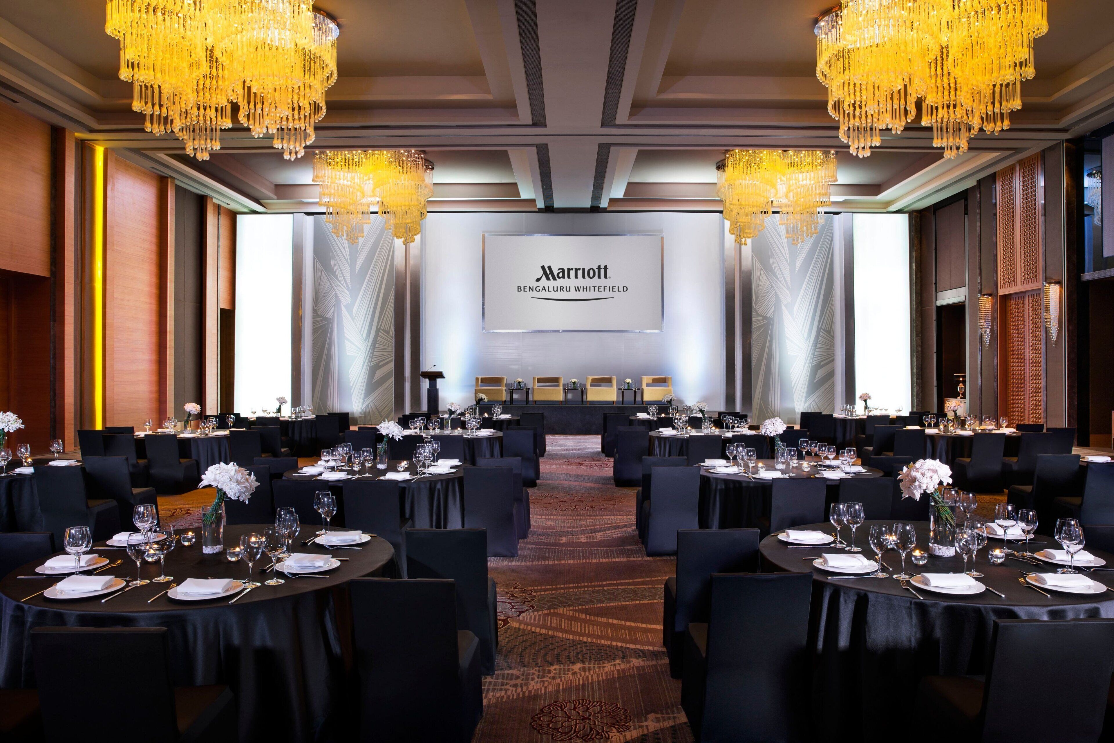 A conference room with tables set for a meeting, chandeliers, and a large screen displaying the Marriott logo.
