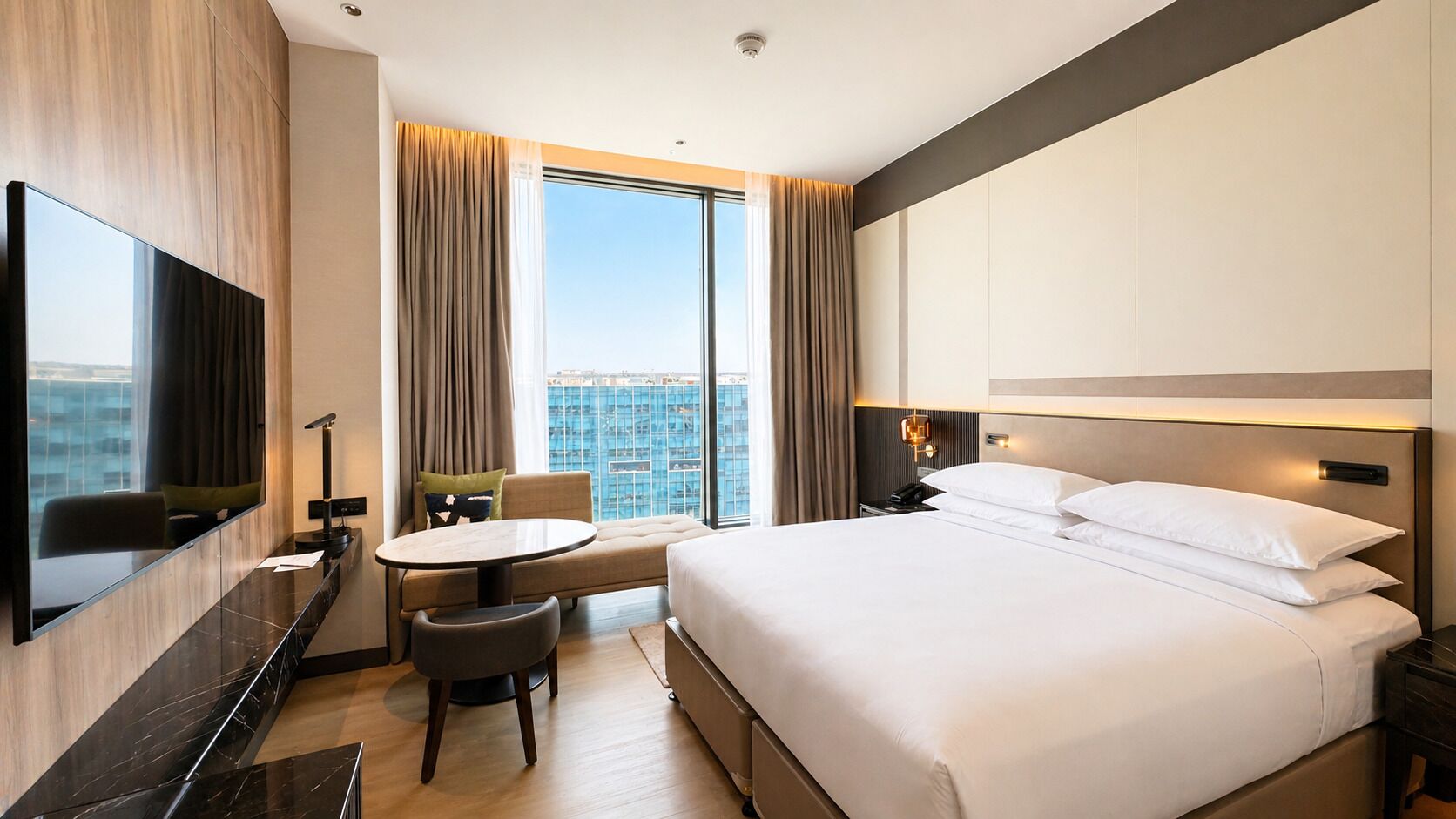 Une chambre d’hôtel avec un lit, un bureau, une chaise, une table ronde et une grande fenêtre avec des rideaux.