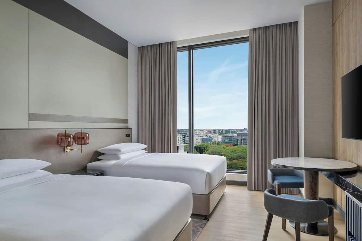 A hotel room with two beds, a small table, a chair, a TV, and a large window with curtains.