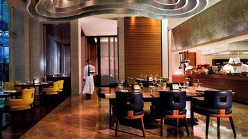 A modern restaurant with a sleek interior, featuring a long dining table with chairs, a bar area, and a view of the cityscape through large windows.