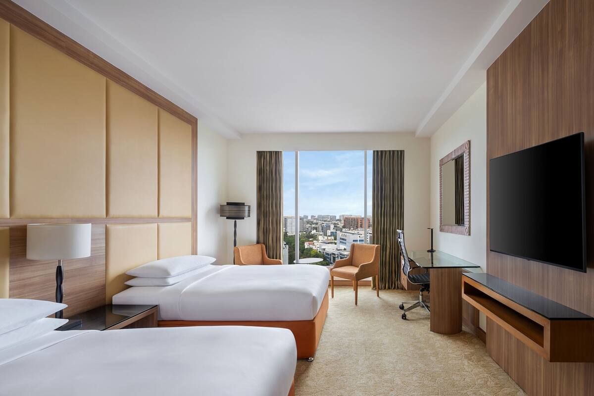 A hotel room with two beds, a desk, a chair, a TV, and a city view.