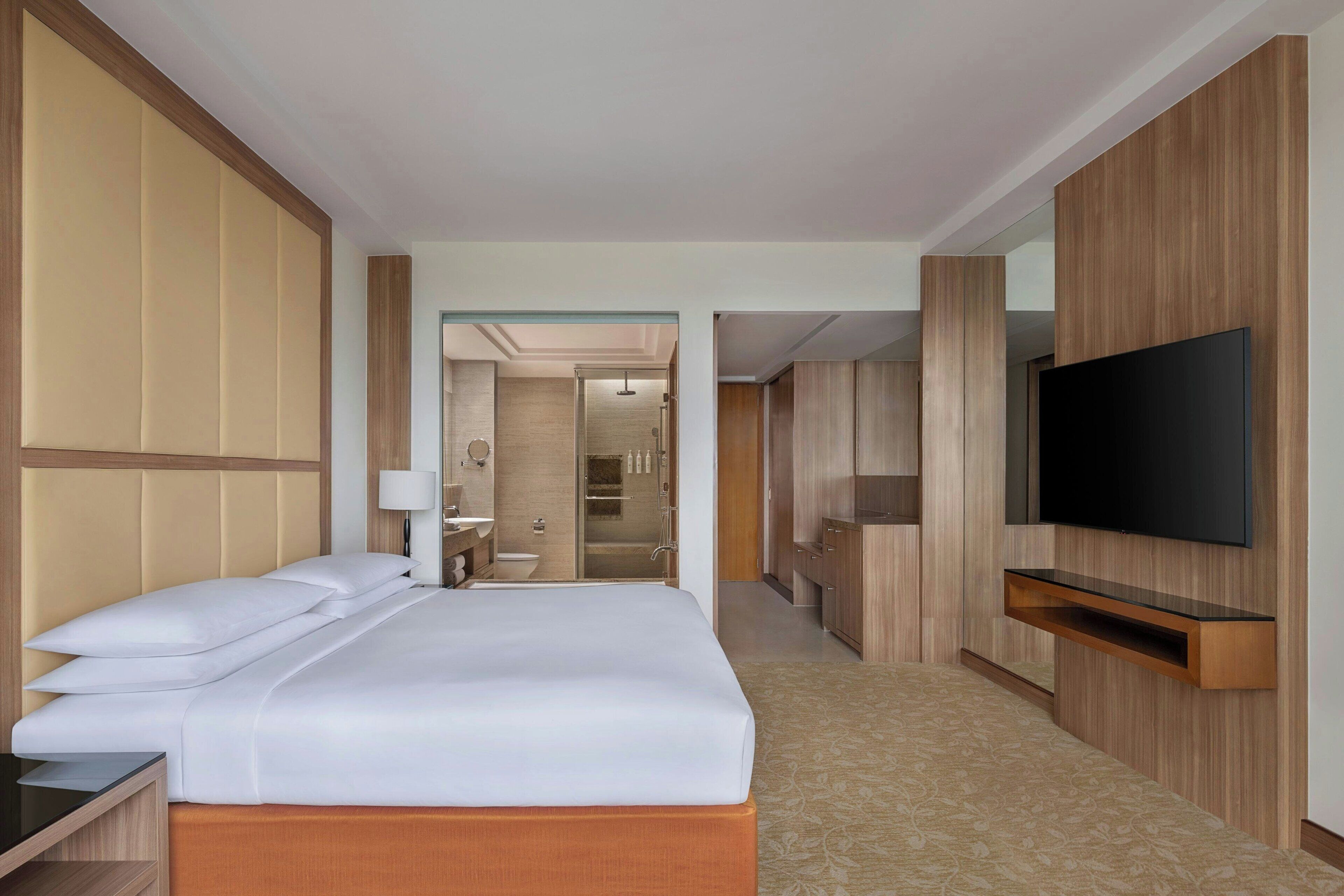 Ein Hotelzimmer mit einem großen Bett, einem an der Wand montierten Fernseher, einem Badezimmer mit Dusche und einem Flur, der zu einem anderen Zimmer führt.