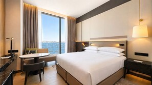 Habitación de hotel con una cama grande, un escritorio con silla, una mesa y vistas a la ciudad a través de amplios ventanales.