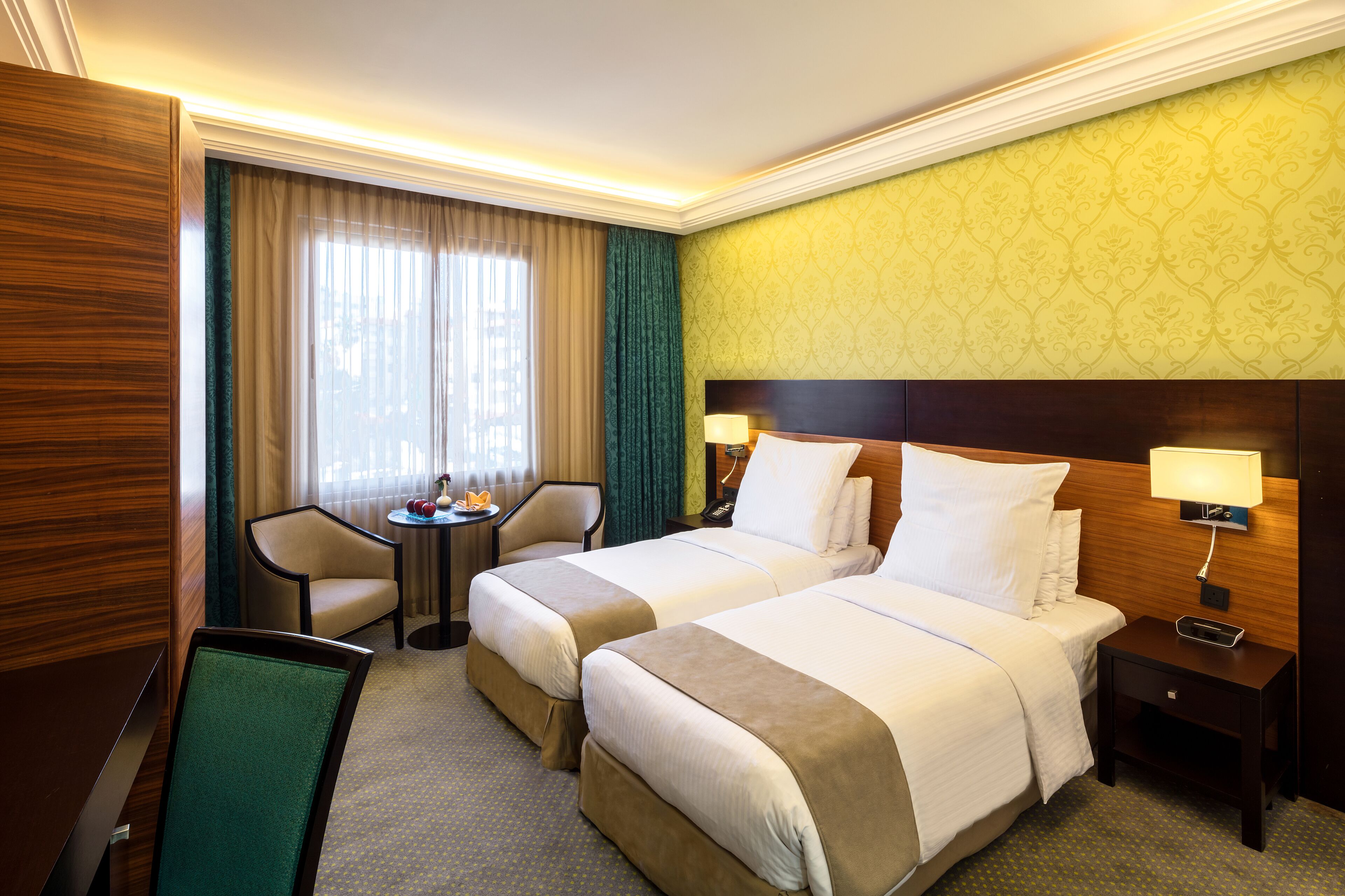 A hotel room with two beds, a desk, a chair, a small table with a lamp, and a window with curtains.