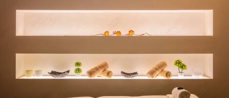 A spa room with a massage table, two illuminated shelves with towels, bowls, and small potted plants.