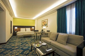 A hotel room with a bed, a sofa, a coffee table with a remote control, a desk with a lamp, and a painting on the wall.