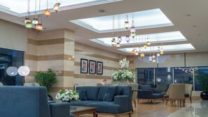 A modern lobby with a high ceiling, skylights, and hanging decorative lights. There are seating areas with sofas and chairs, and a large potted plant. - Corp Amman Hotel (Amman)