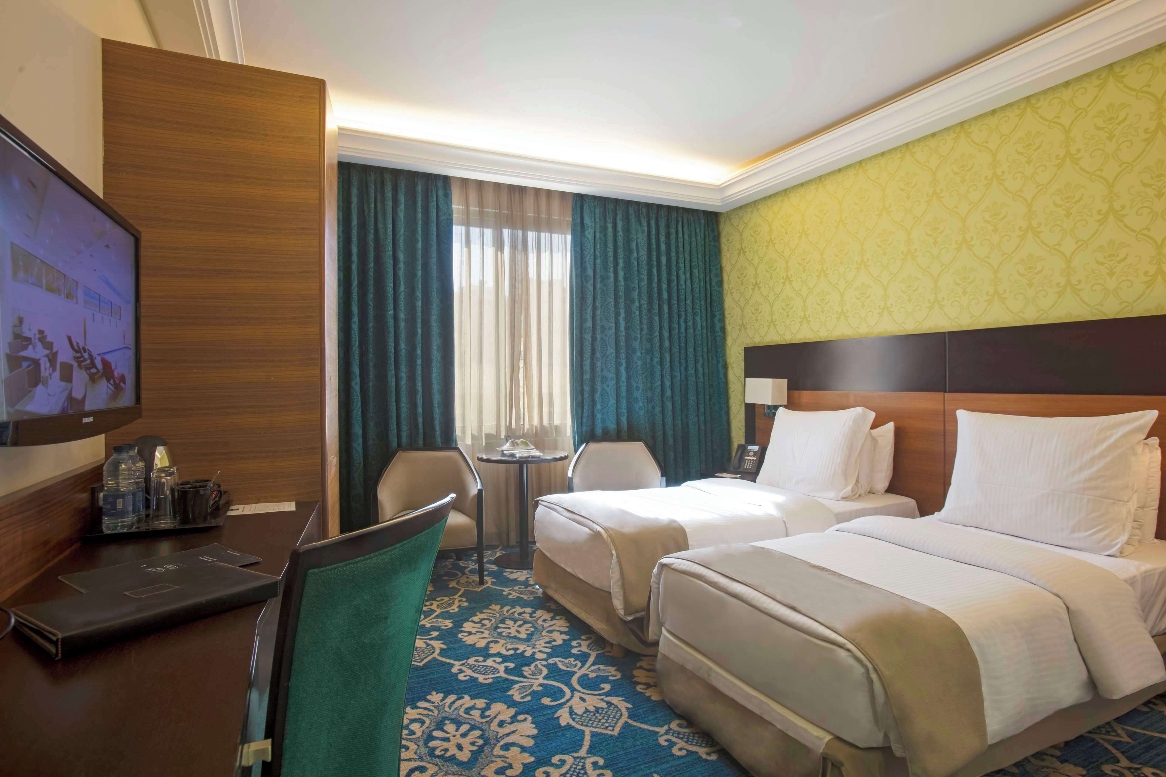 A hotel room with two beds, a desk with a laptop, a TV, and a small table with a lamp.