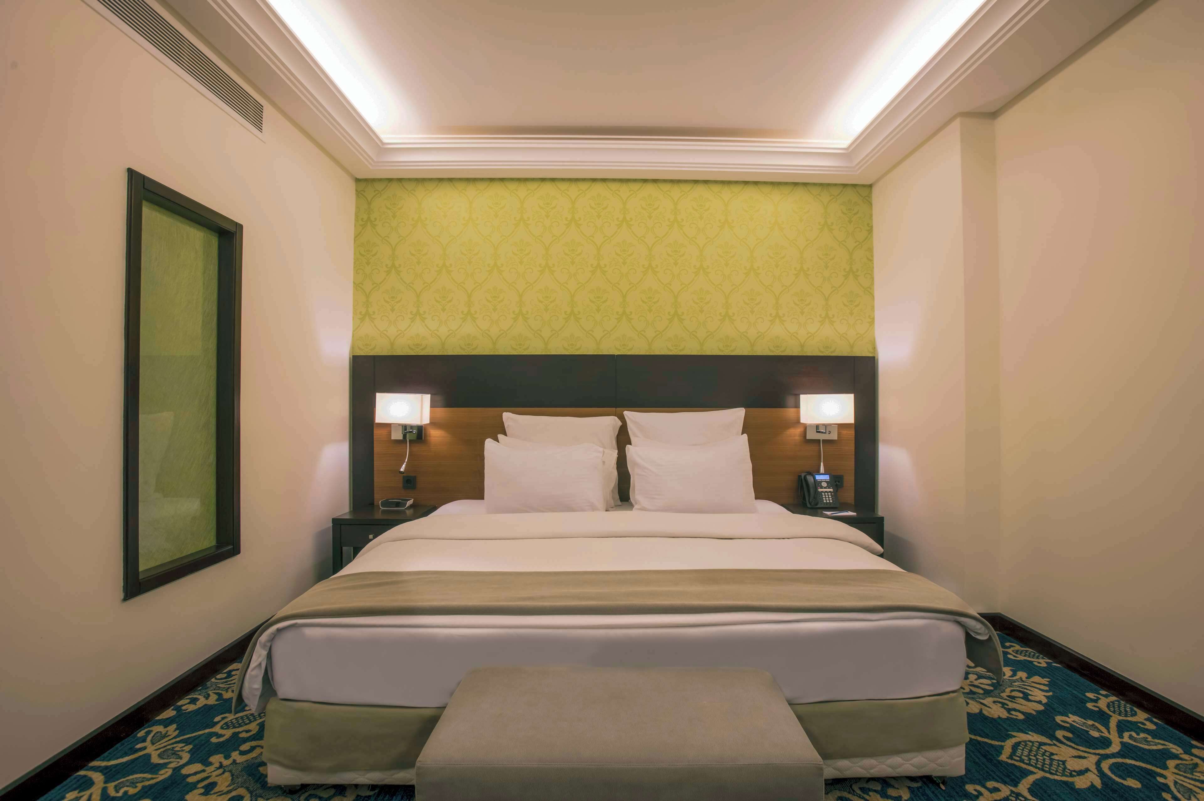 Une chambre d’hôtel avec un grand lit, deux tables de chevet avec des lampes, une tête de lit, un tableau encadré au mur et une moquette à motifs.