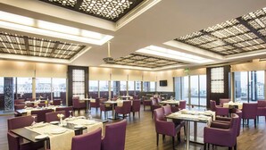 A spacious restaurant with multiple tables and chairs, large windows offering city views, and a modern ceiling design. - Corp Amman Hotel (Amman)