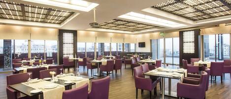 A spacious restaurant with multiple tables and chairs, large windows offering city views, and a modern ceiling design.