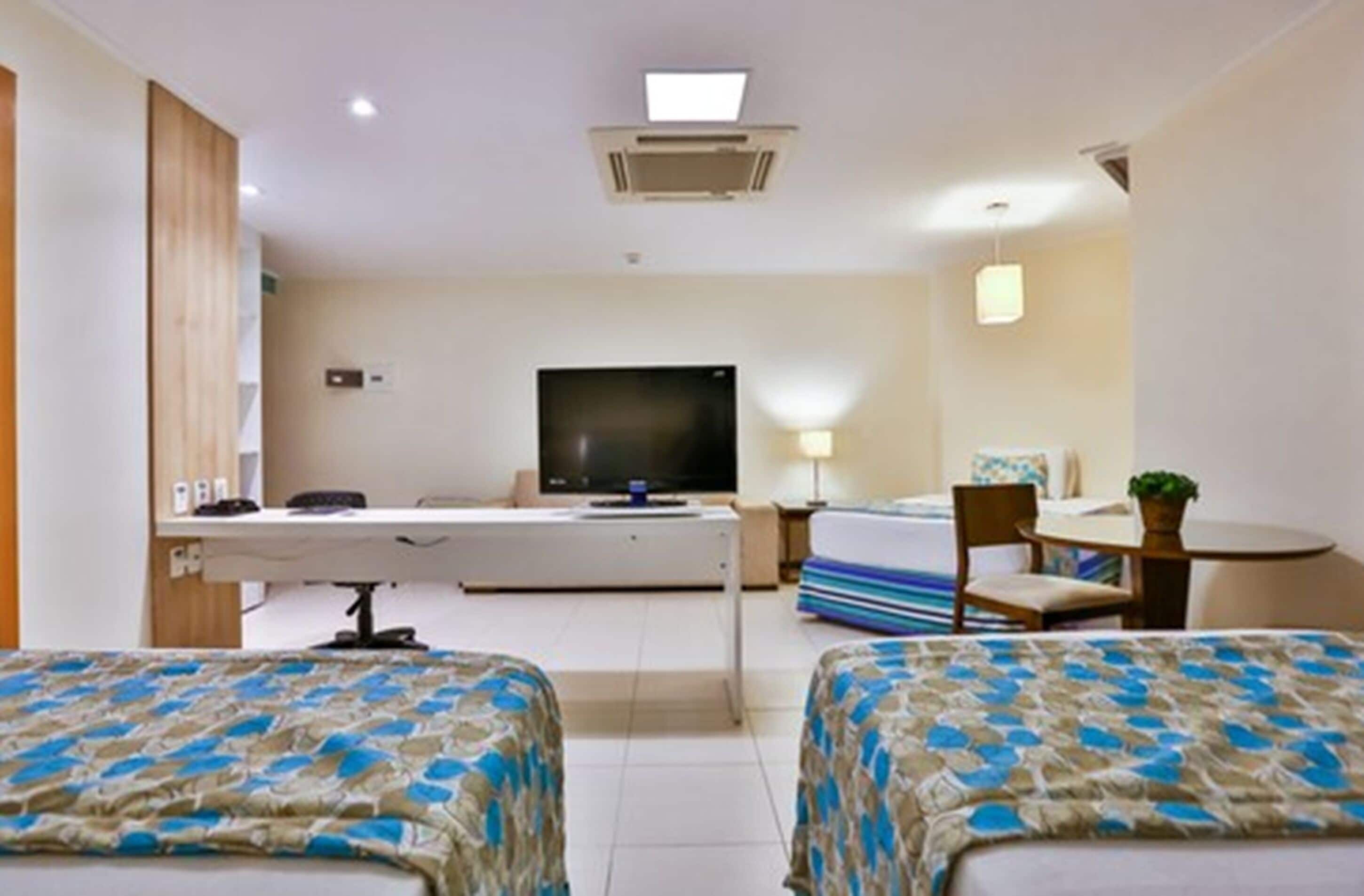 A hotel room with two beds, a desk with a computer, a flat-screen TV, a small table with a chair, and a bedside lamp.