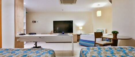 A hotel room with two beds, a desk with a computer, a flat-screen TV, a small table with a chair, and a bedside lamp.