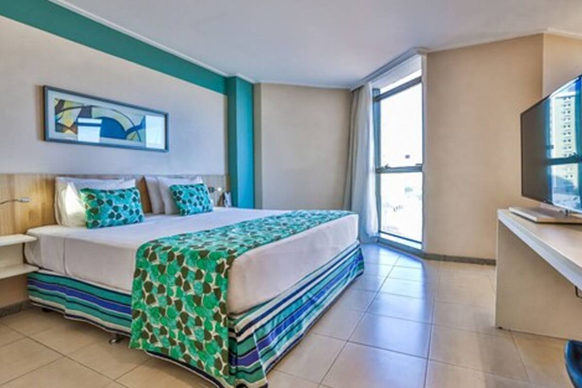 Quarto de hotel com cama, escrivaninha, televisão e janela.
