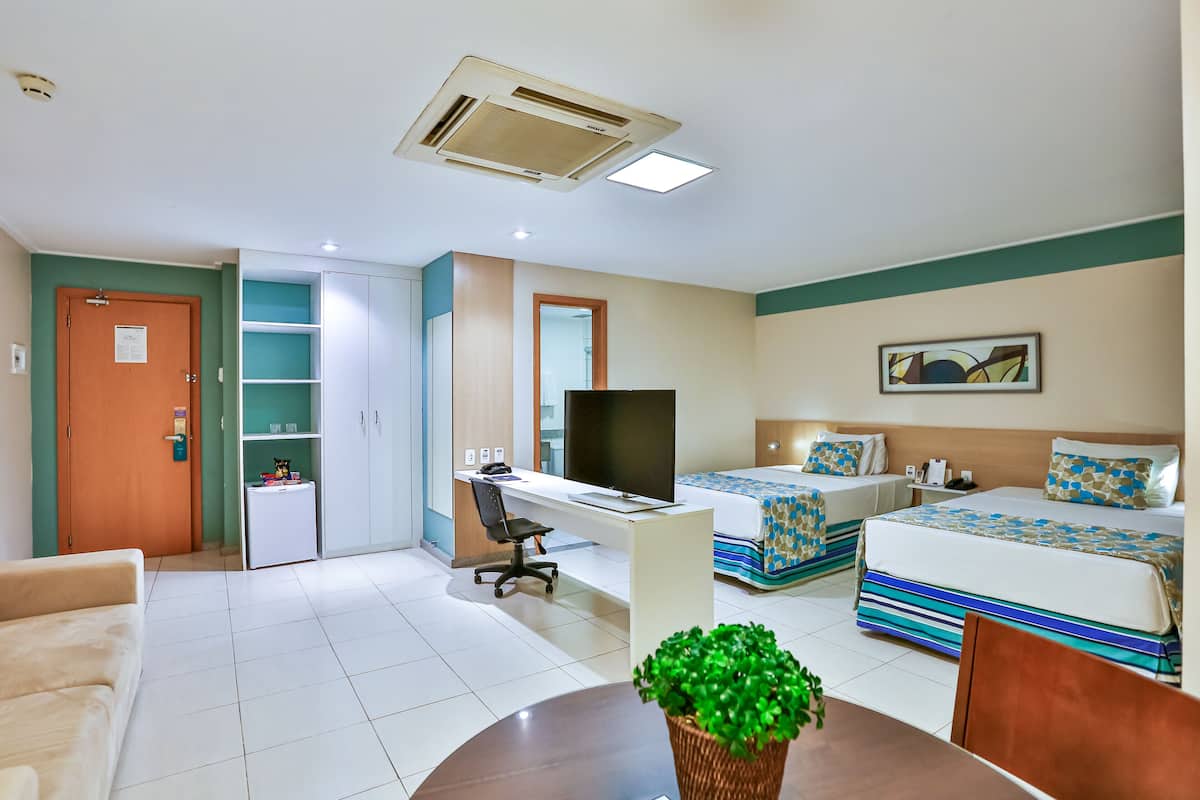 Quarto de hotel com duas camas, uma escrivaninha, uma cadeira, uma televisão, uma mesinha de centro com uma planta, e banheiro com chuveiro e vaso sanitário.