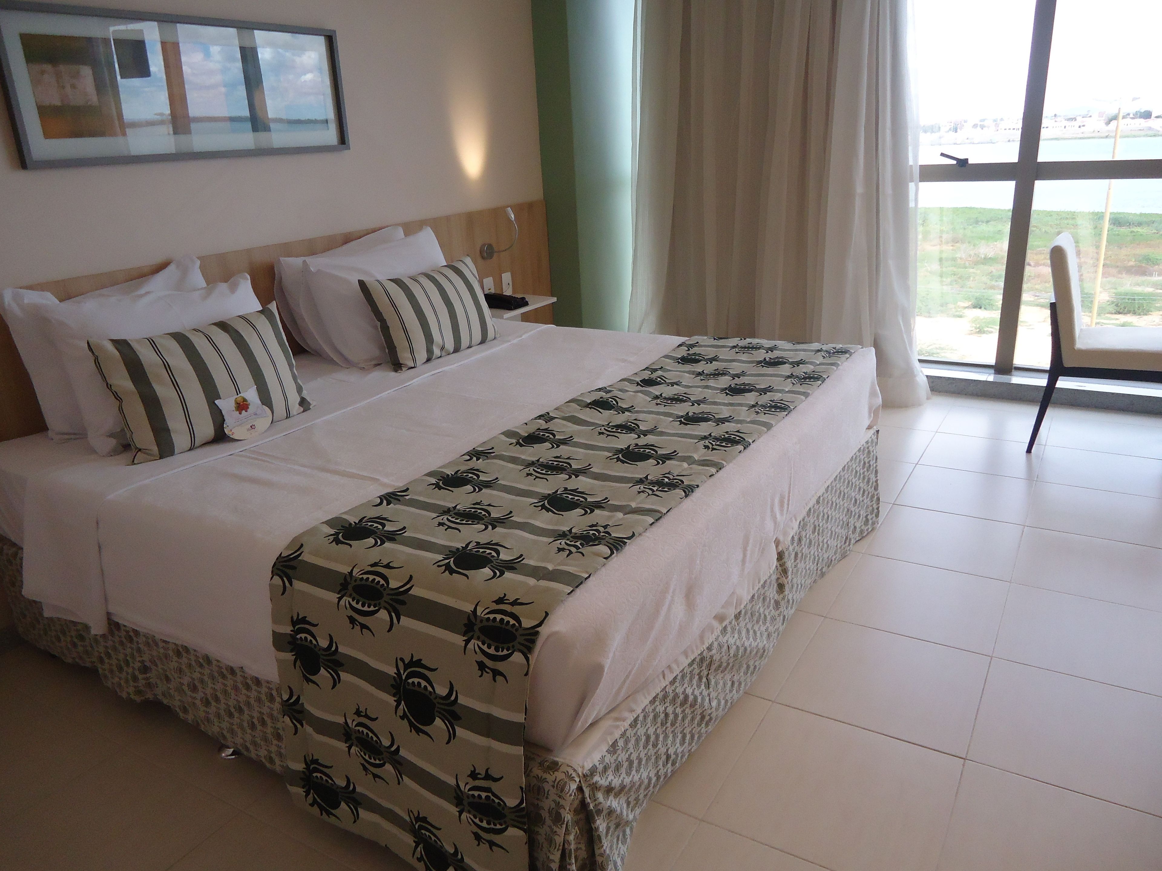 Una cama bien hecha con un almohadón a rayas y una colcha estampada, una mesita de noche con una lámpara, y vistas al exterior a través de un ventanal grande.