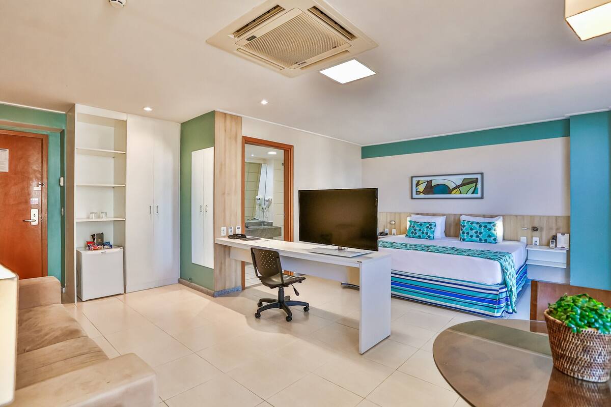 Quarto de hotel moderno com cama, escrivaninha, cadeira, televisão e uma pequena cozinha.
