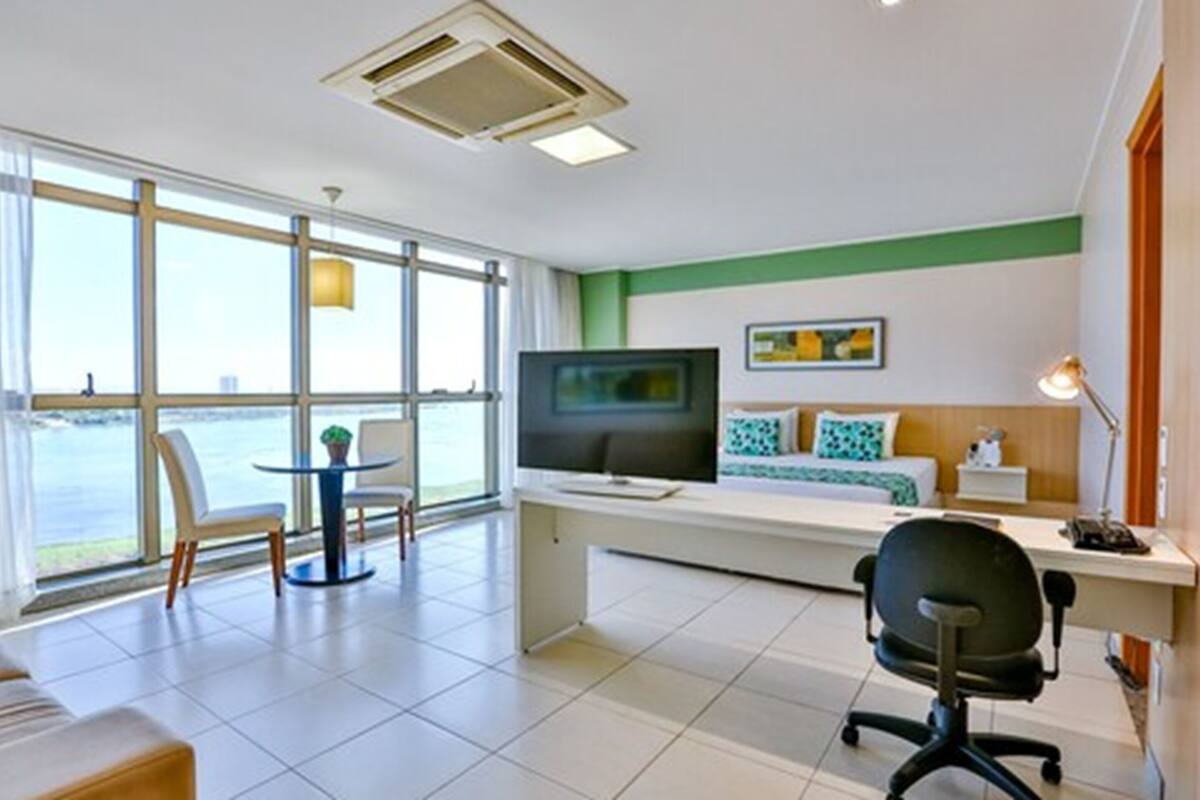 Quarto de hotel moderno com uma janela grande, uma escrivaninha, uma cama e uma televisão.