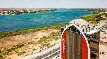 Un edificio moderno con fachada roja y blanca, cerca de una masa de agua, con una autopista y un área urbana al fondo.