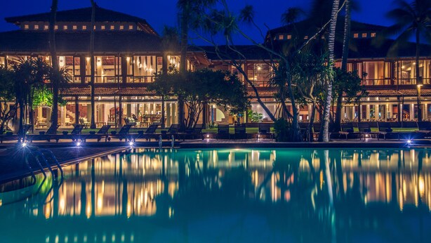 A well-lit resort with a swimming pool, palm trees, and a large building with multiple windows.