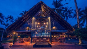 A thatched-roof structure with a glass enclosure, surrounded by palm trees and outdoor seating.