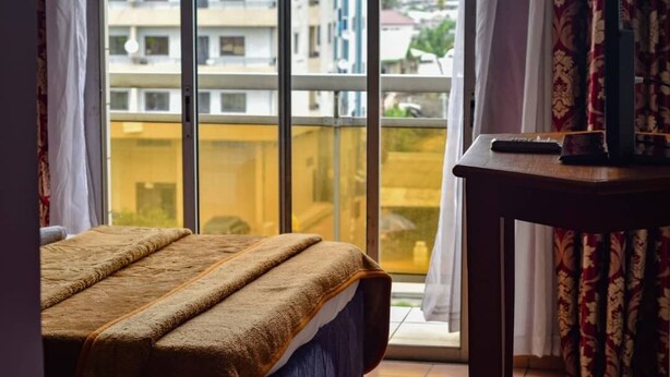 Ein Schlafzimmer mit einem großen Fenster, einem Bett mit brauner Decke, einem Holztisch und einer Fernbedienung.