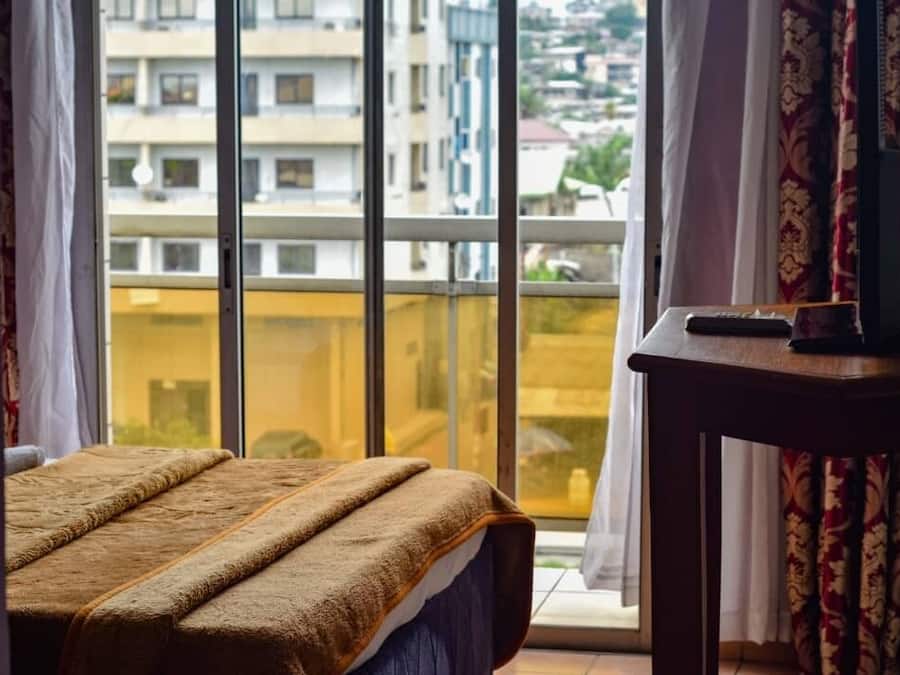 A bedroom with a large window, a bed with a brown blanket, a wooden table, and a remote control.