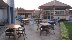 Un patio al aire libre con mesas y sillas, un pequeño edificio con techo de tejas y un jardín con seto.